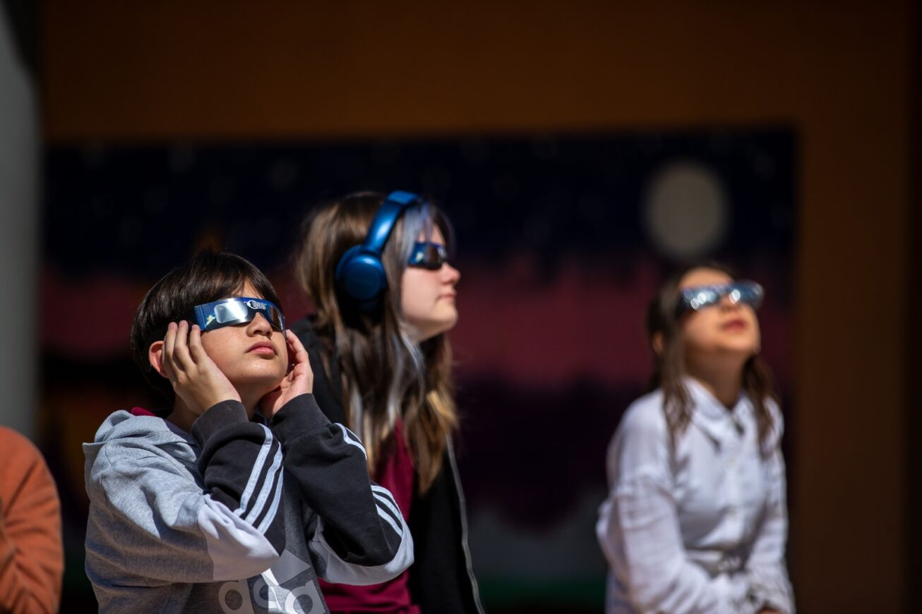 Three students stand outside looking at the eclipse with their special glasses